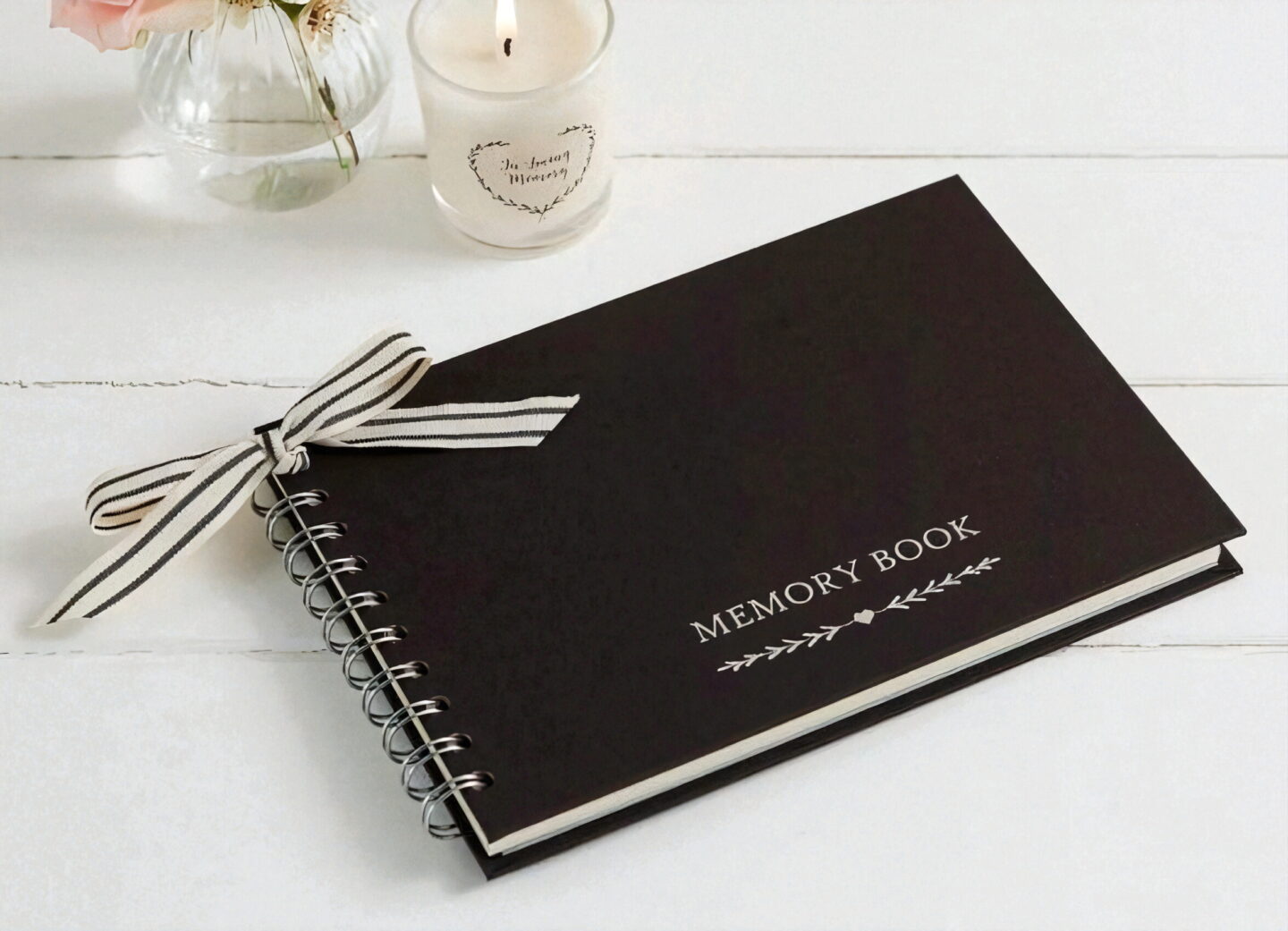 A black memory book with a white striped ribbon tied in a bow sits on a white wooden surface, the words 'Memory Book' printed on the cover.